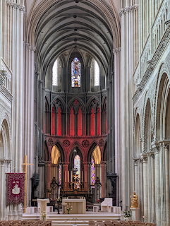 Gothic cathedral interior with ribbed vaulted ceiling, stone columns, red altar backdrop, and stained glass