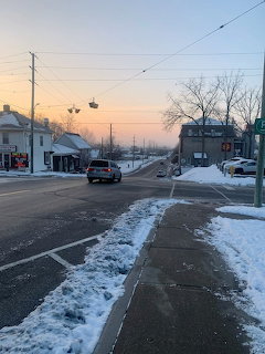Snowy sidewalk beside a small-town road at sunset with cars, bare trees, and utility poles