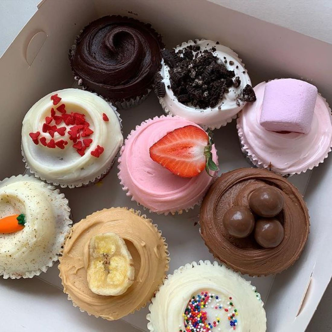 Overhead view of nine decorated cupcakes with chocolate, vanilla, strawberry, and other toppings in a white box