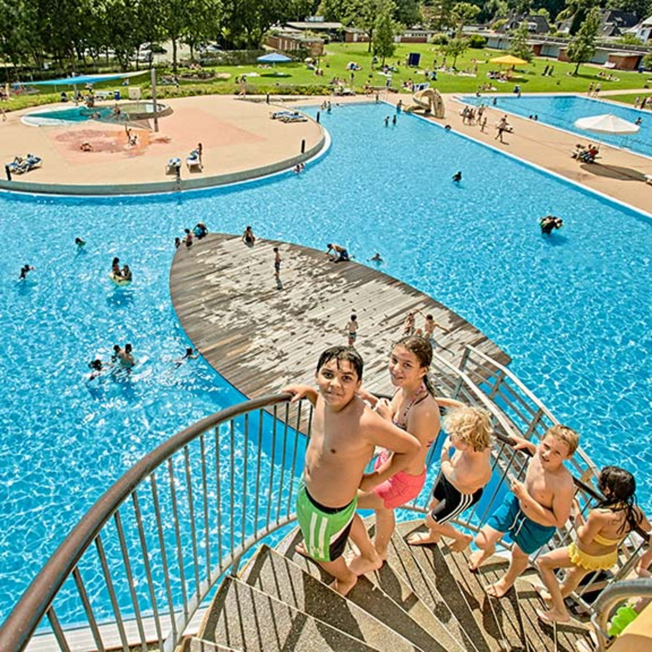 Children on spiral pool steps beside a large blue outdoor swimming pool