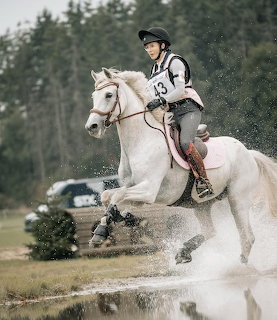 Rider in helmet and numbered vest on white horse jumping through water during cross-country eventing
