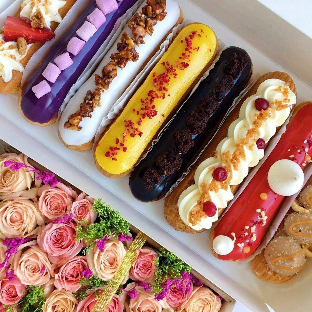 Five decorated eclairs in purple, yellow, brown, white, and red glazes arranged on white serving tray with pink roses