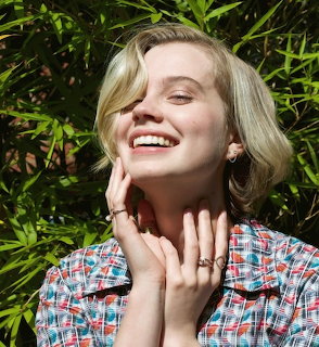 Young blonde woman smiling with hand near face, wearing plaid shirt, green leafy background