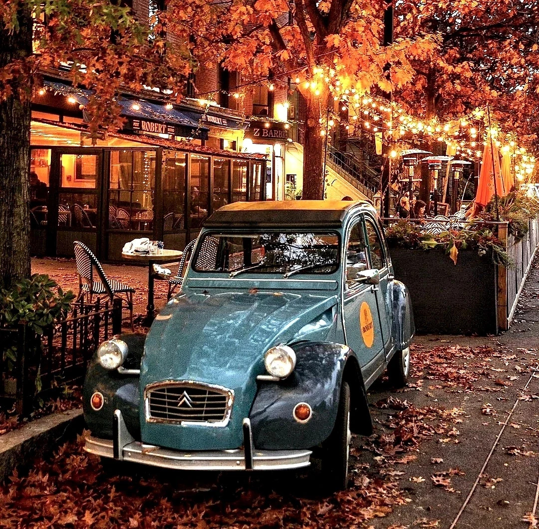 Blue vintage Citroen 2CV car on cobblestone street with cafes and fall foliage at dusk