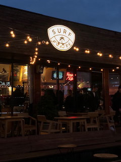 Warmly lit cafe exterior at dusk with string lights, wooden tables, and a round sign