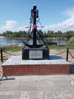 Black anchor monument on brick base beside river with boats and trees in background