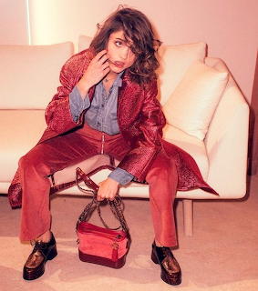 Woman in red sequined jacket sitting on beige chair, hand near face, studio setting