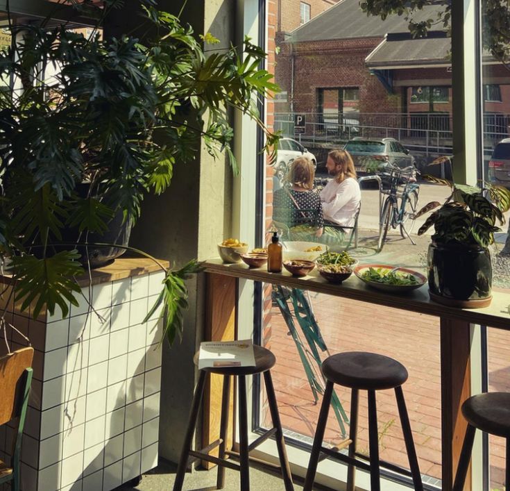 Bright cafe interior with plants, wooden bar stools, and large windows overlooking a city street