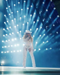 Female performer in a white cowboy hat and sparkly outfit on a concert stage with bright blue lights