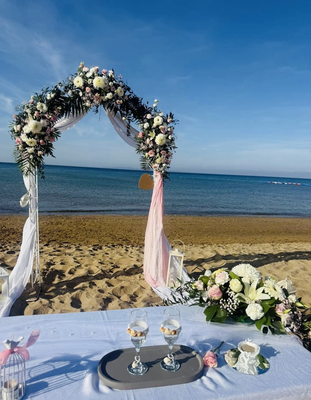 White flower-adorned wedding arch with flowing fabric on beach, ocean and blue sky behind, table with flowers in front