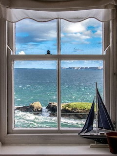 View through a white window frame of a rocky coastline with blue-green sea, cliffs, and a model sailboat on theill