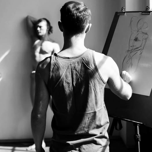 Black-and-white photo of a person drawing at an easel with a blurred shirtless model behind