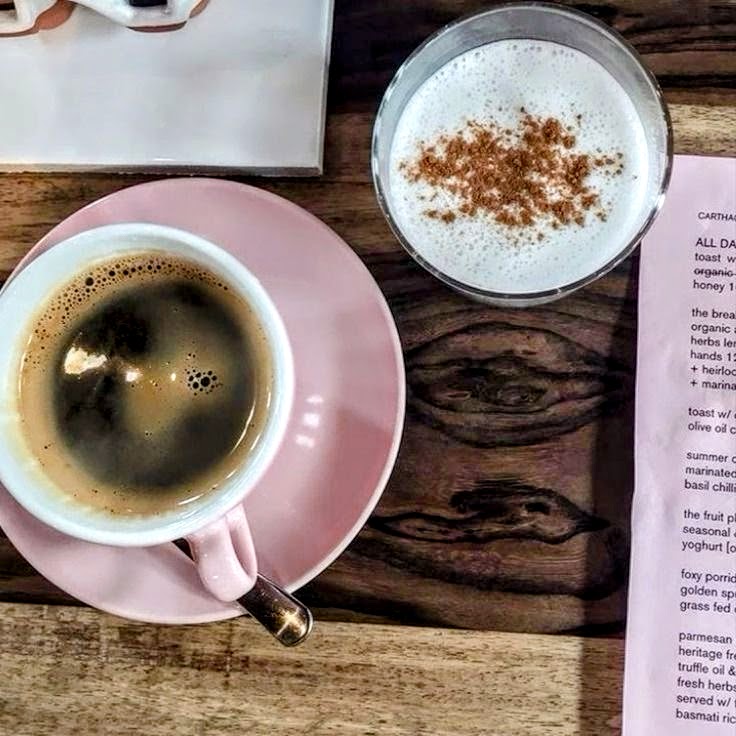 Black coffee in pink cup beside cappuccino with cinnamon sprinkled on top, coffee beans scattered on wooden table