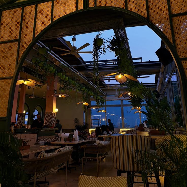 Cozy cafe interior with tables, chairs, plants, and a large arched opening at dusk