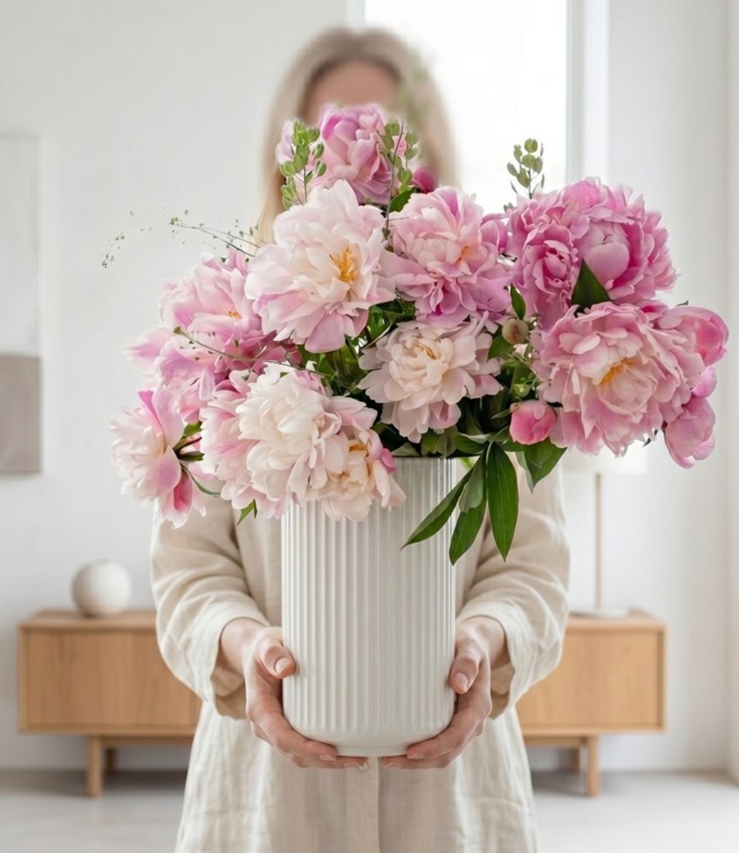 Person holding a white vase of pink peonies indoors