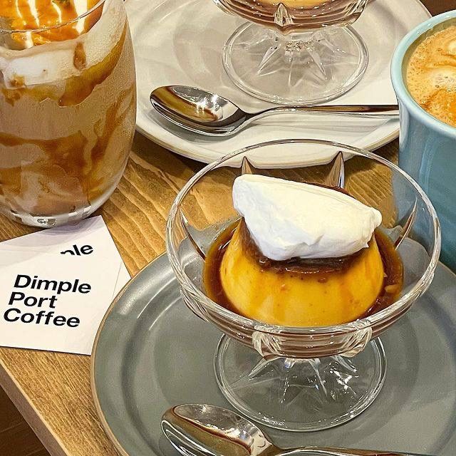 Caramel pudding with whipped cream in a glass dish beside iced and hot coffee on a table