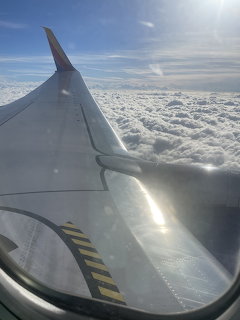 Airplane wing seen from a passenger window above white clouds and blue sky