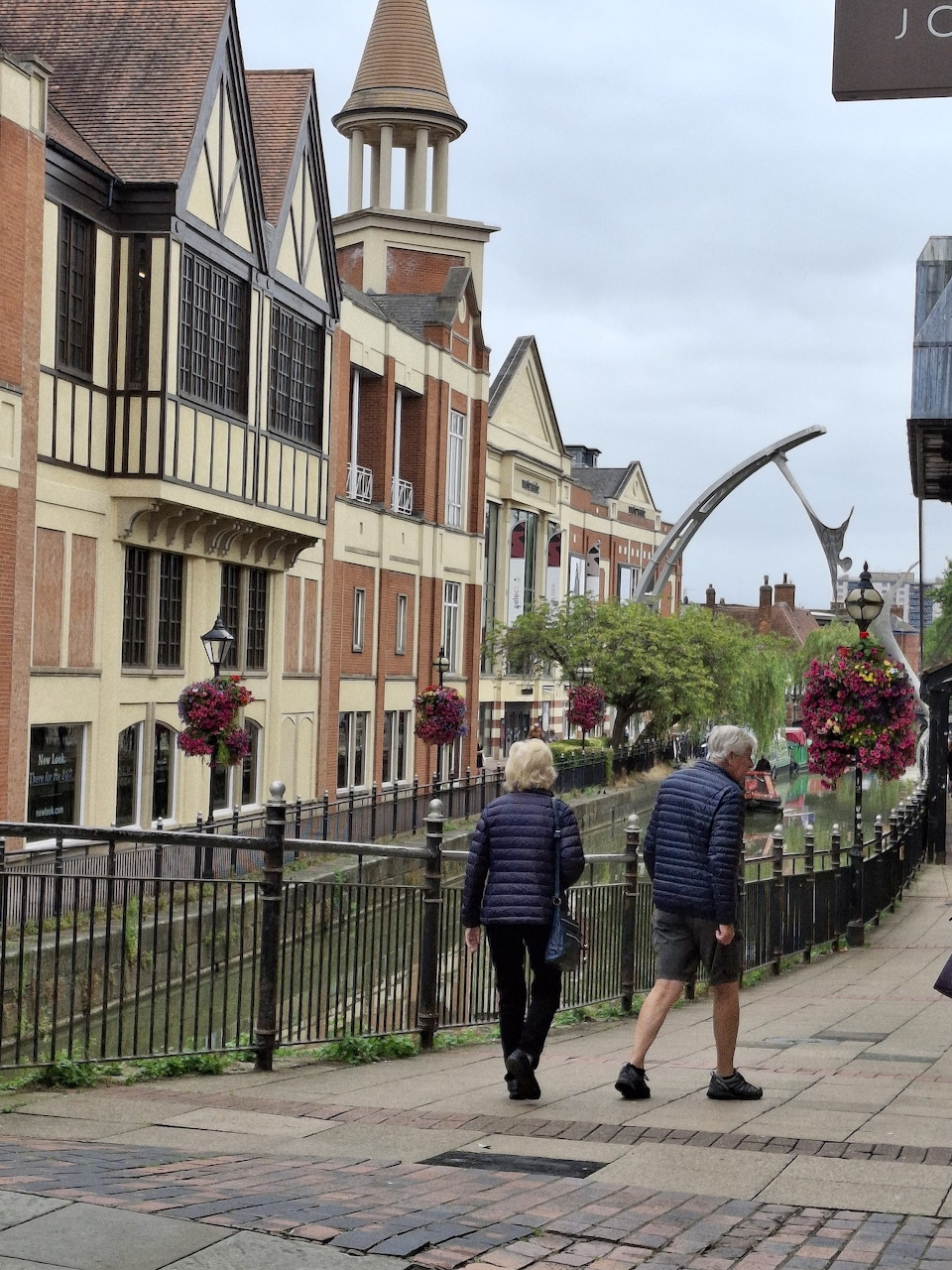 Two pedestrians walking along a path past Tudor-style buildings with hanging flower baskets in an English town