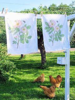 Chickens beneath floral-painted linens hanging on a clothesline in a green farmyard