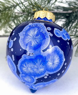 Blue ceramic Christmas ornament with painted white snowflakes and gold top hanging near pine branches