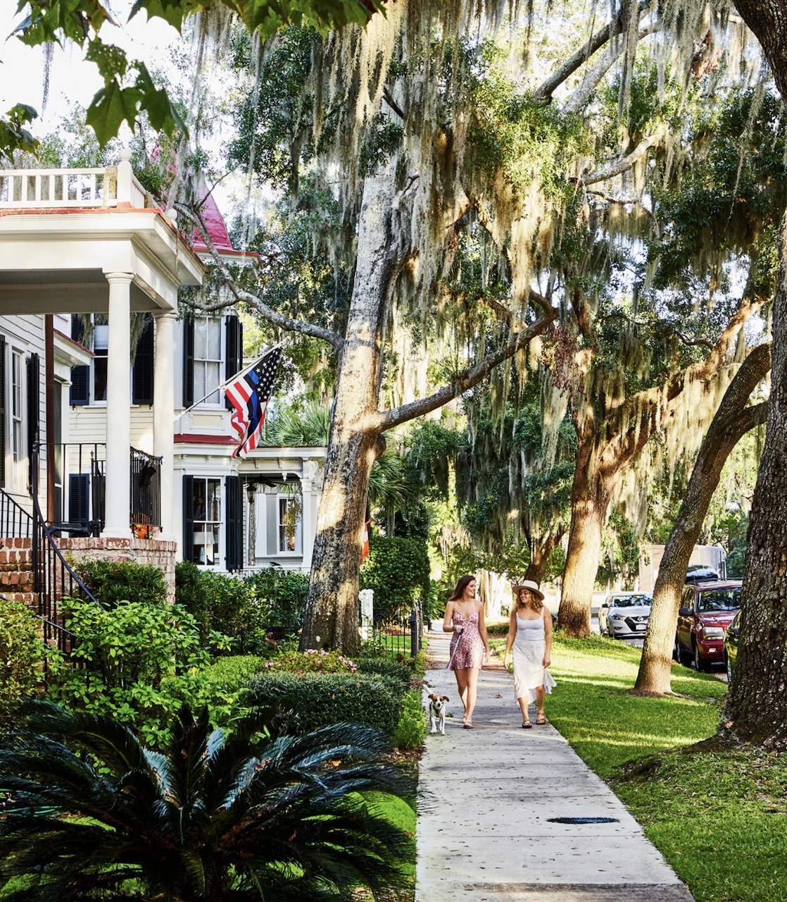Two women walking on a sidewalk beside white houses and large moss-covered trees
