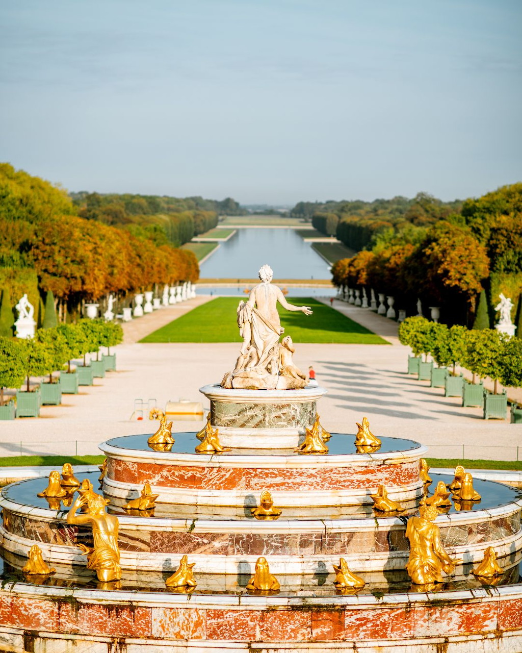 Golden fountain sculpture in manicured gardens with tree-lined path extending to distant canal under overcast sky