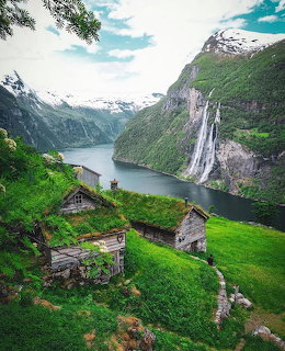 Rustic cottages on a green hillside above a fjord with mountains and a tall waterfall