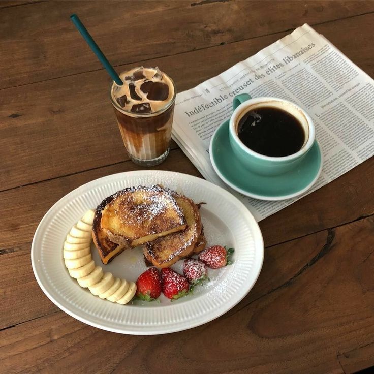 Breakfast plate with French toast, banana slices, berries, coffee cup, and iced beverage on wooden table