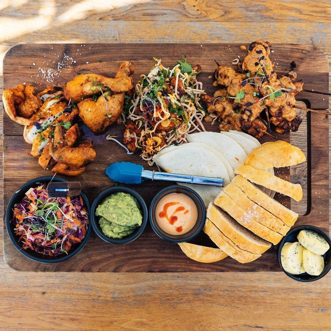 Overhead view of Asian fusion chicken platter with fried items, noodles, breads, and colorful sauces on rustic wooden board