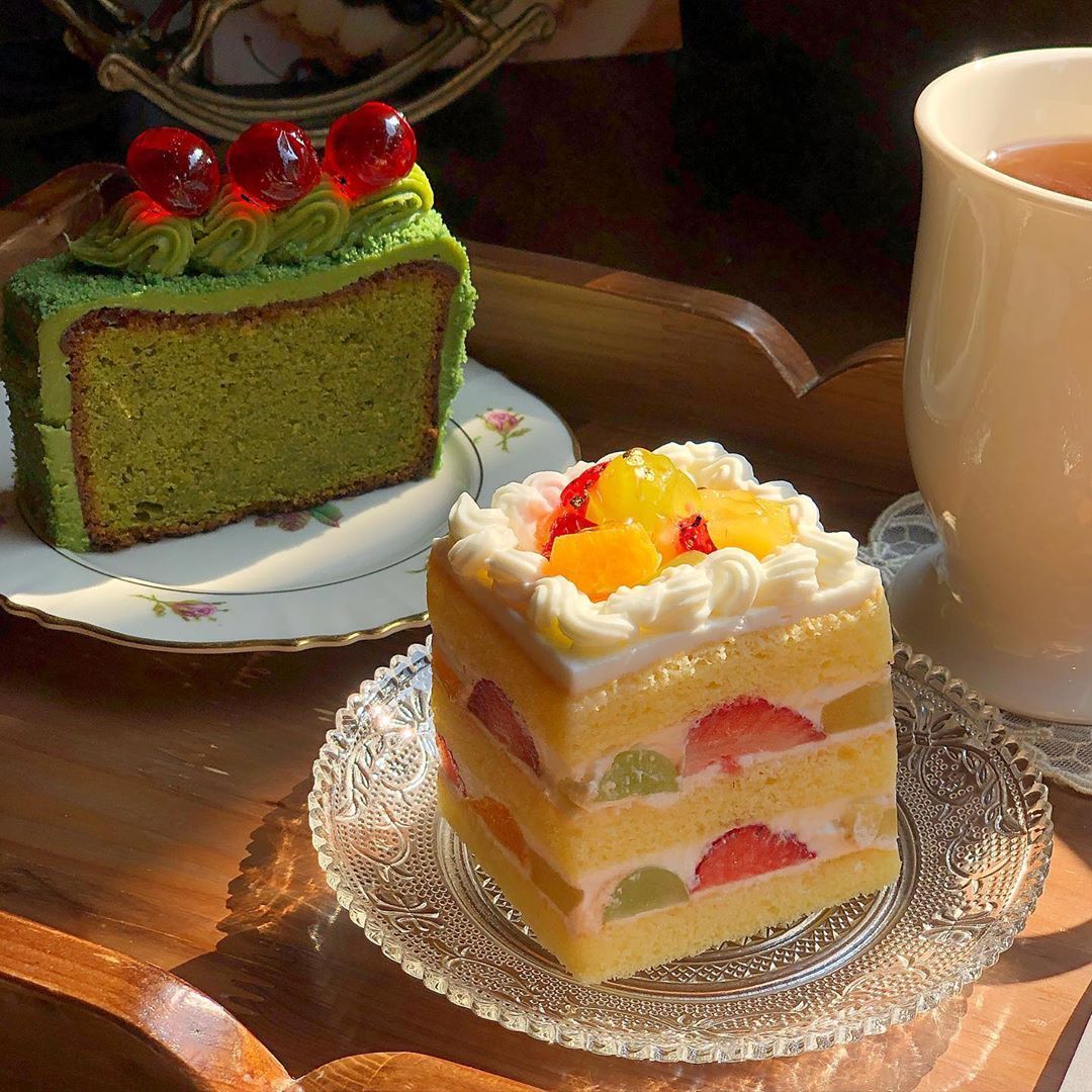 Two slices of cake on plates beside a cup of tea in warm sunlight