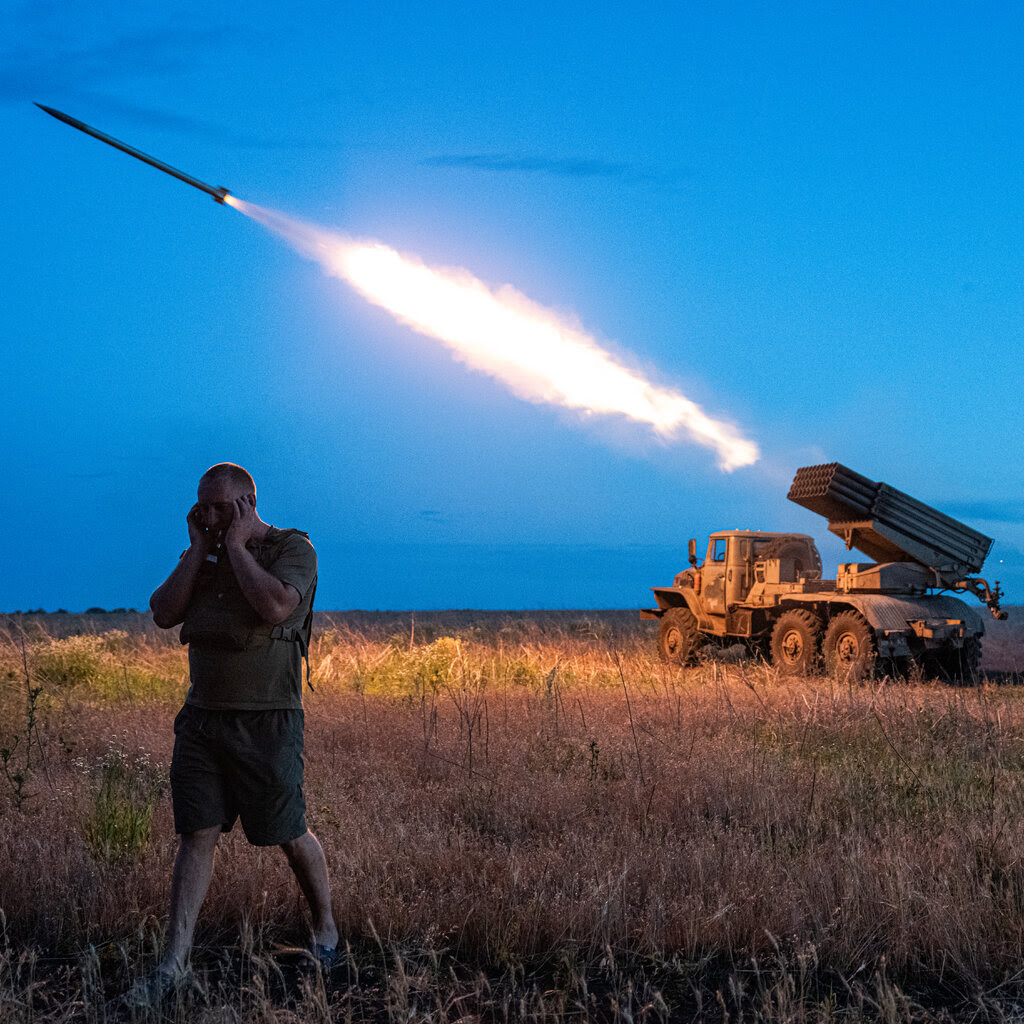 Man covering his ears as a rocket launches from a military truck in a field at dusk
