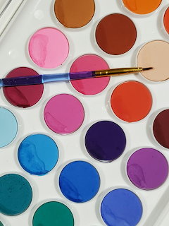 White watercolor palette with colorful circular paint wells and a paintbrush resting on top