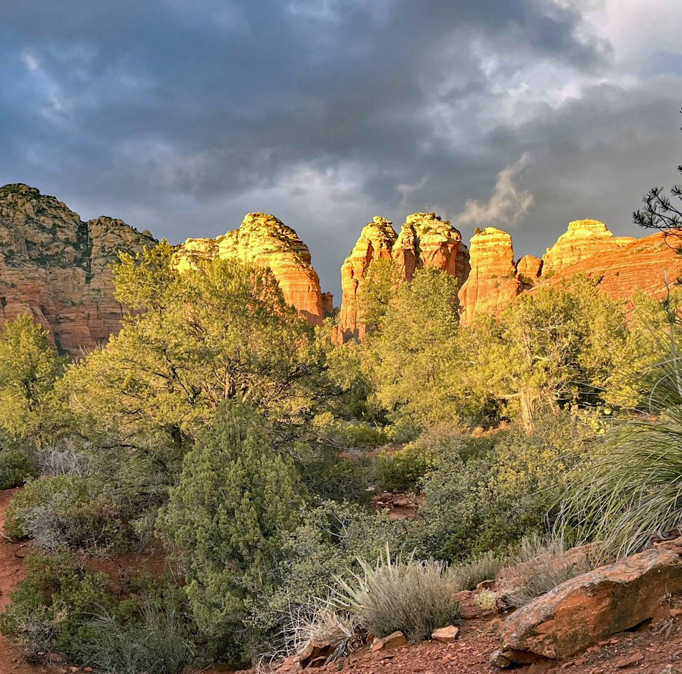 Sunlit red rock formations towering over green desert vegetation beneath cloudy sky