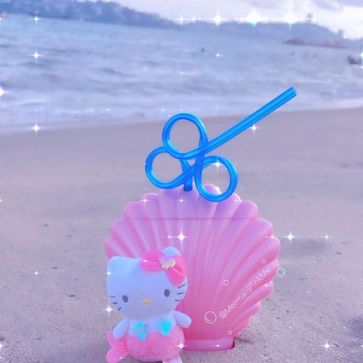 Pink Hello Kitty shell cup with blue straw on a sandy beach near the ocean