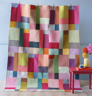 Colorful patchwork quilt hanging on wall with multicolored rectangular fabric patches and red side table