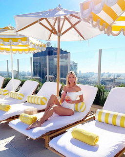 Blonde woman sitting on white lounge chair at rooftop pool with yellow towels and umbrellas, city skyline behind
