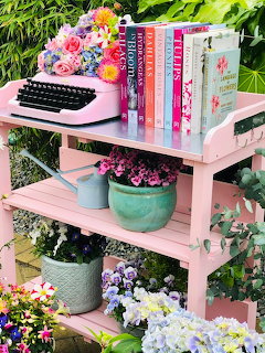 Pink garden cart with vintage typewriter, books, and flower pots surrounded by blooming hydrangeas