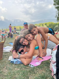 Mother and two daughters hugging on grass near a lake with mountains in the background