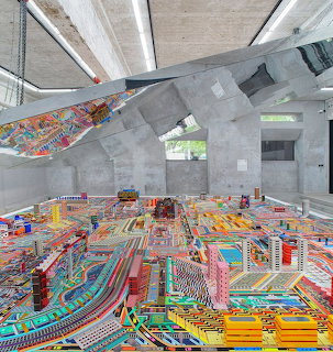 Colorful miniature city art installation covering the floor of a modern concrete gallery with angular roof