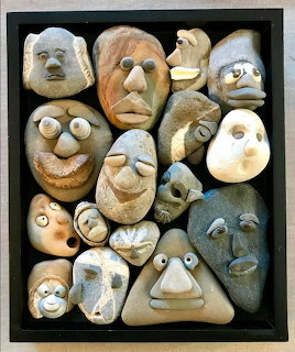 Collection of rocks painted as expressive human faces arranged in a black shadow box frame