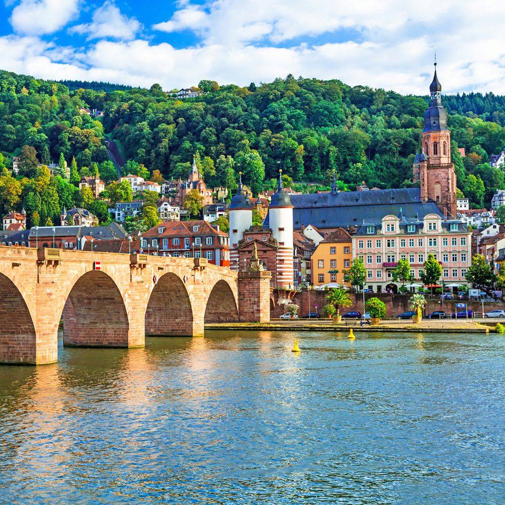 European riverside village with arched stone bridge, red church tower, and green forested hills