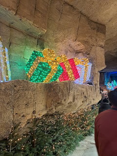 Colorful lit gift box decorations on a stone cave wall with Christmas trees below at an underground market