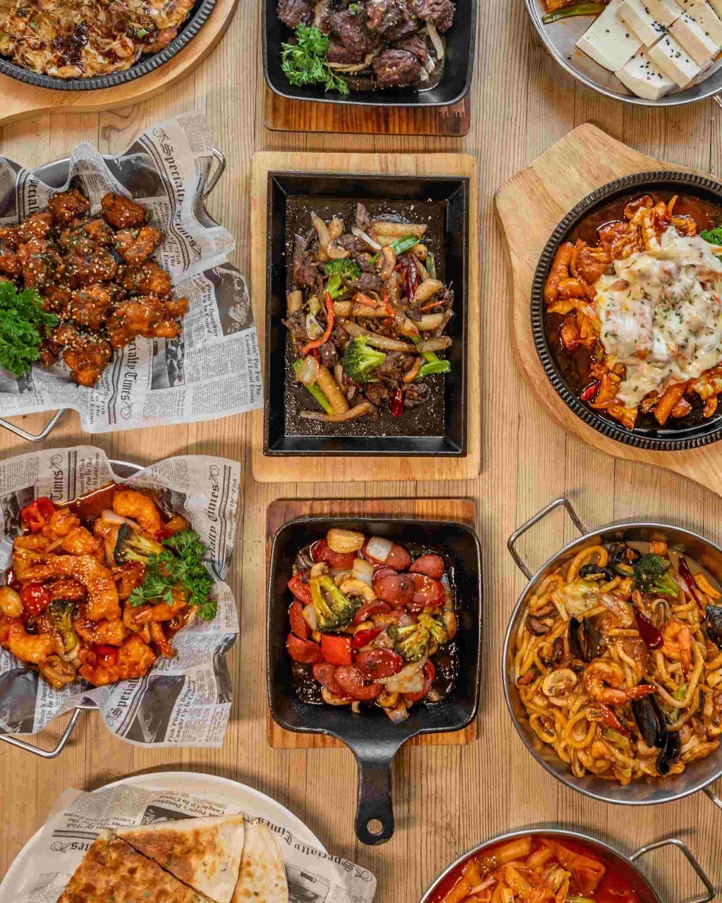 Top-down view of various Asian dishes including stir-fries, noodles, and seafood in pans and bowls on a wooden table.