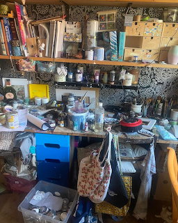 Cluttered artist workspace with shelves full of paint jars, brushes, bottles, and craft supplies