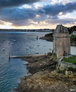 Stone tower on rocky coastline overlooking harbor with boats and dramatic cloudy sky