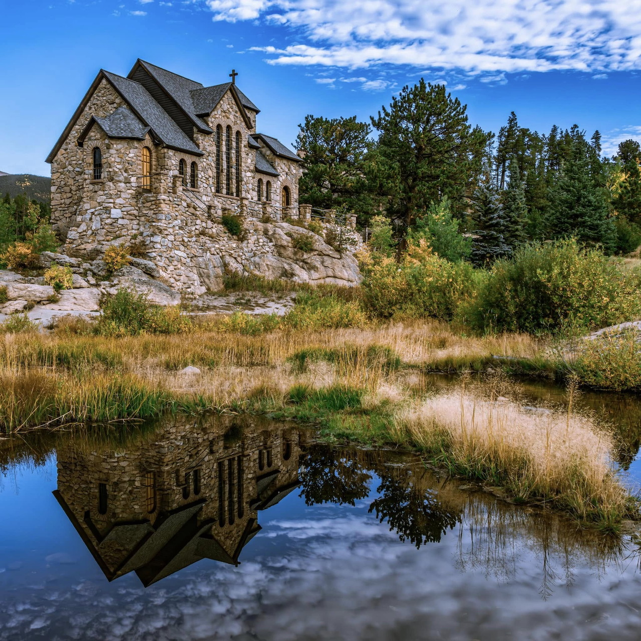 Stone church with reflection in lake, surrounded by trees and mountains under blue sky