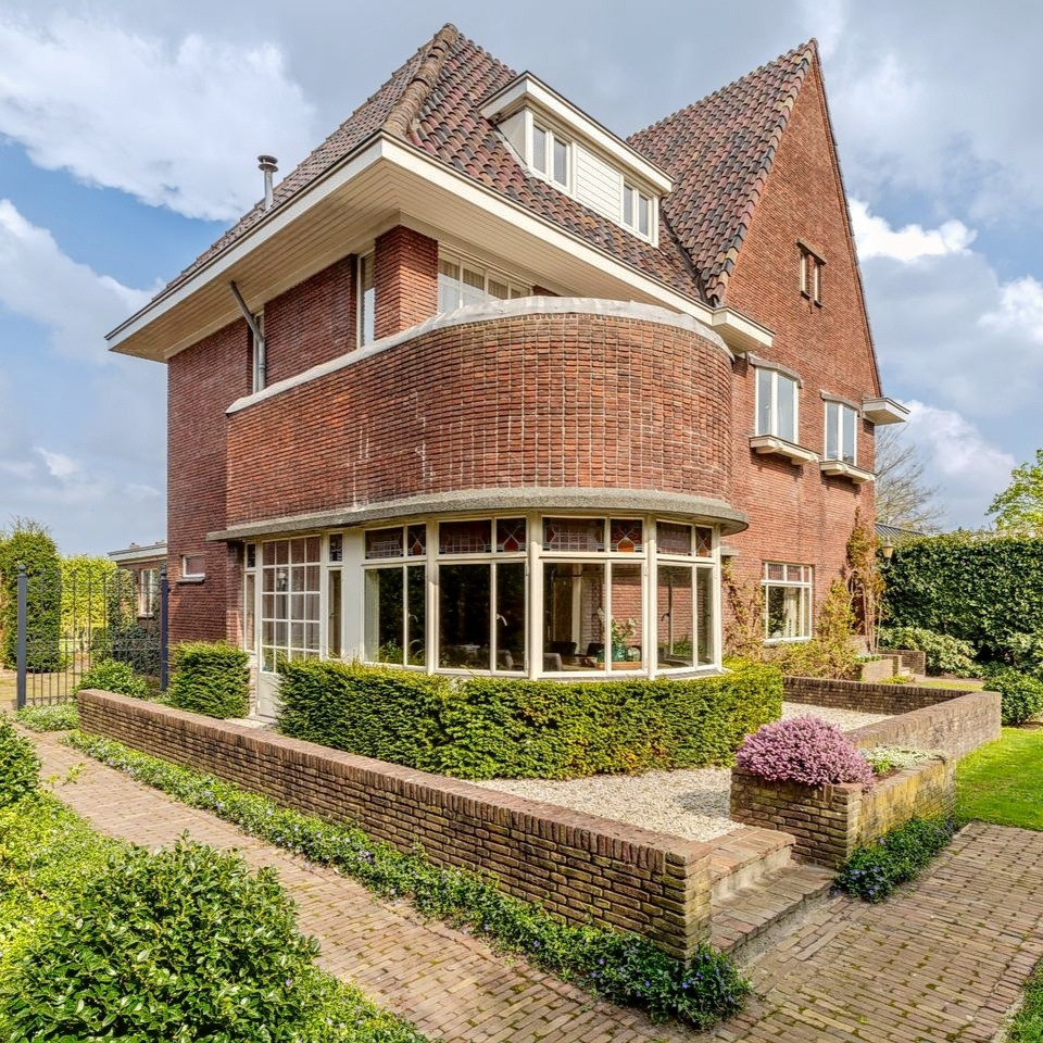 Red brick Art Deco house with curved corner, tiled roof, and landscaped garden with hedges and flowers