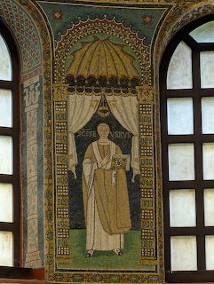 Byzantine mosaic of robed saint figure in ornate gold arch between two windows with intricate geometric patterns