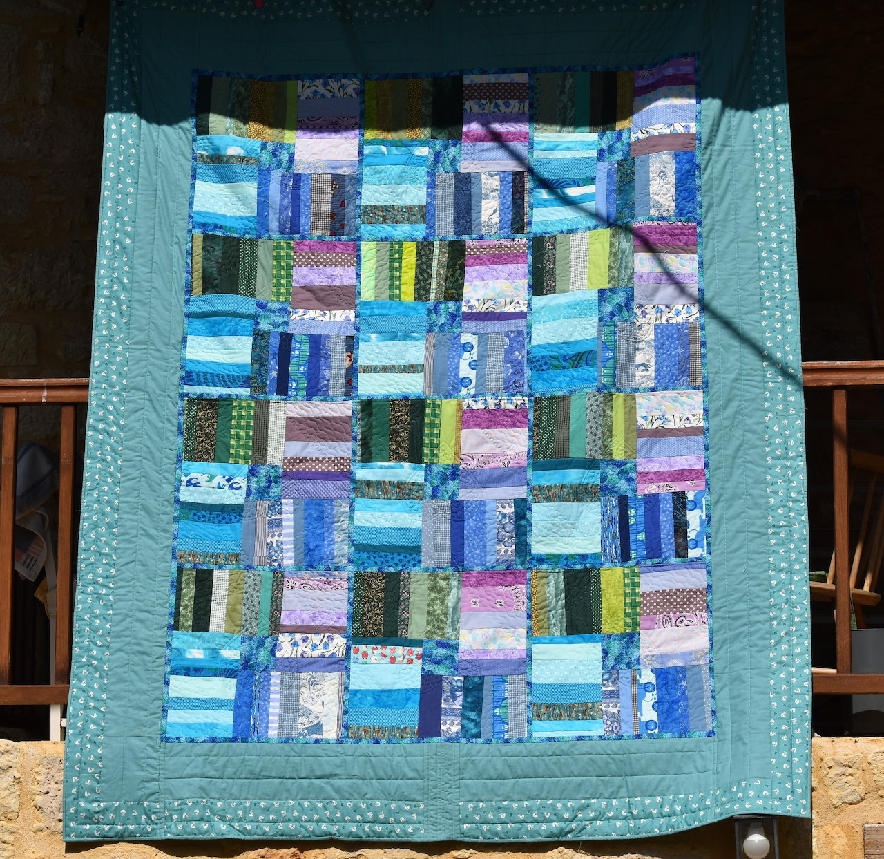 Colorful patchwork quilt with turquoise border hanging on wooden fence outdoors