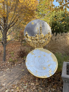 Gold circular sister city marker plaques on a stand surrounded by autumn trees and fallen leaves